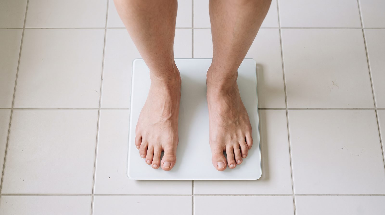person looking tired while standing on a bathroom scale in a plain home bathroom, neutral lifestyle scene